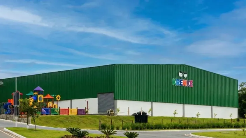 Vista externa de um grande armazém industrial com fachada verde e logotipo “KRENKE”, mostrando estrutura ampla de galpão, área paisagística e céu azul ao fundo
